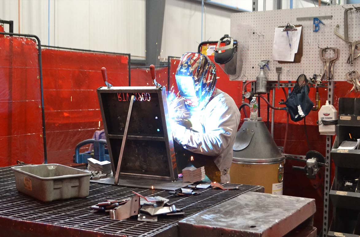 Multi-Faceted Welding A welder works at a metal grate table and welds a piece of metal secured to a black fixture labeled "RTV-X 900"