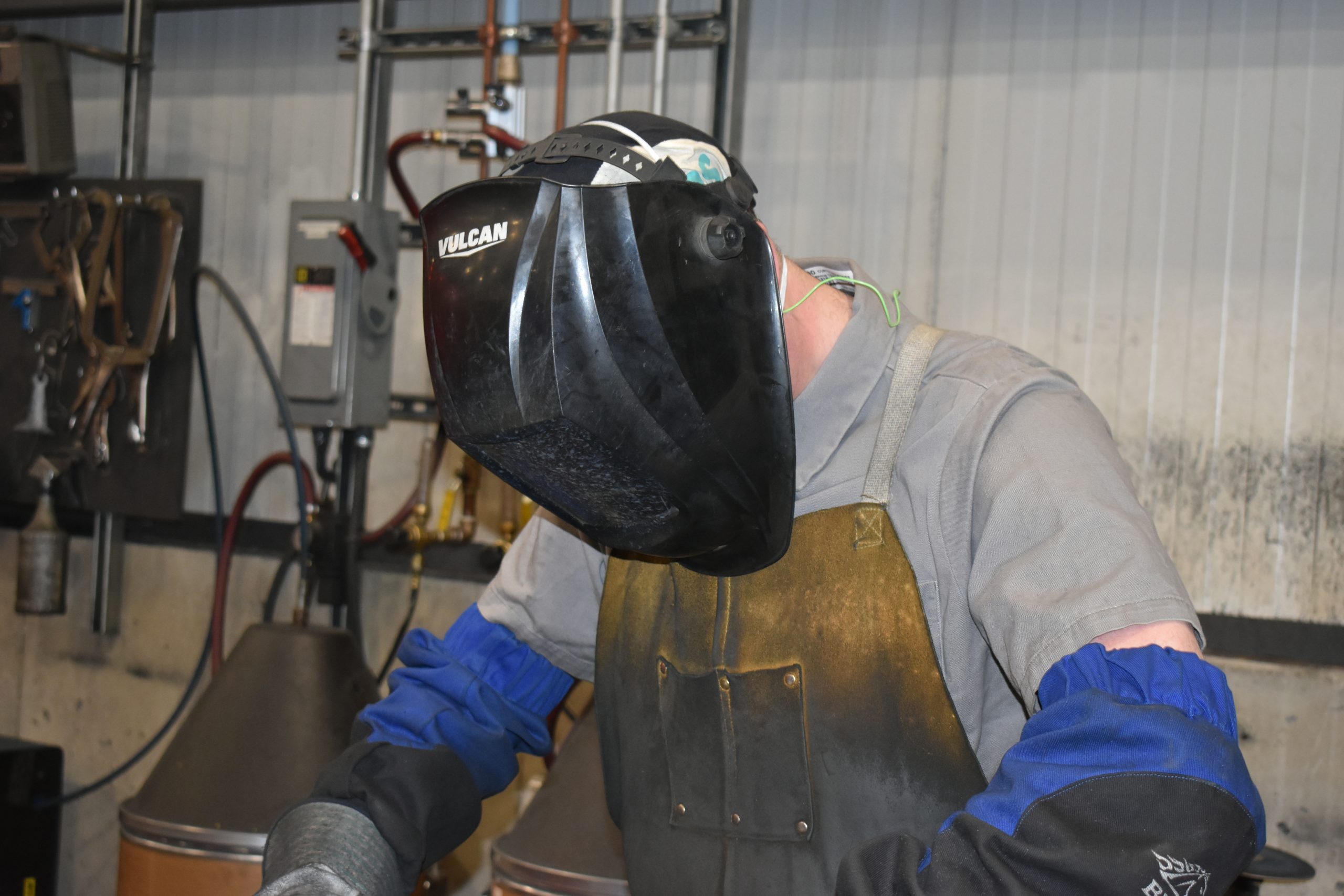 Worker wearing a welding helmet, heavy protective gloves, and a leather apron while performing metalwork inside Curtis Industries' facility