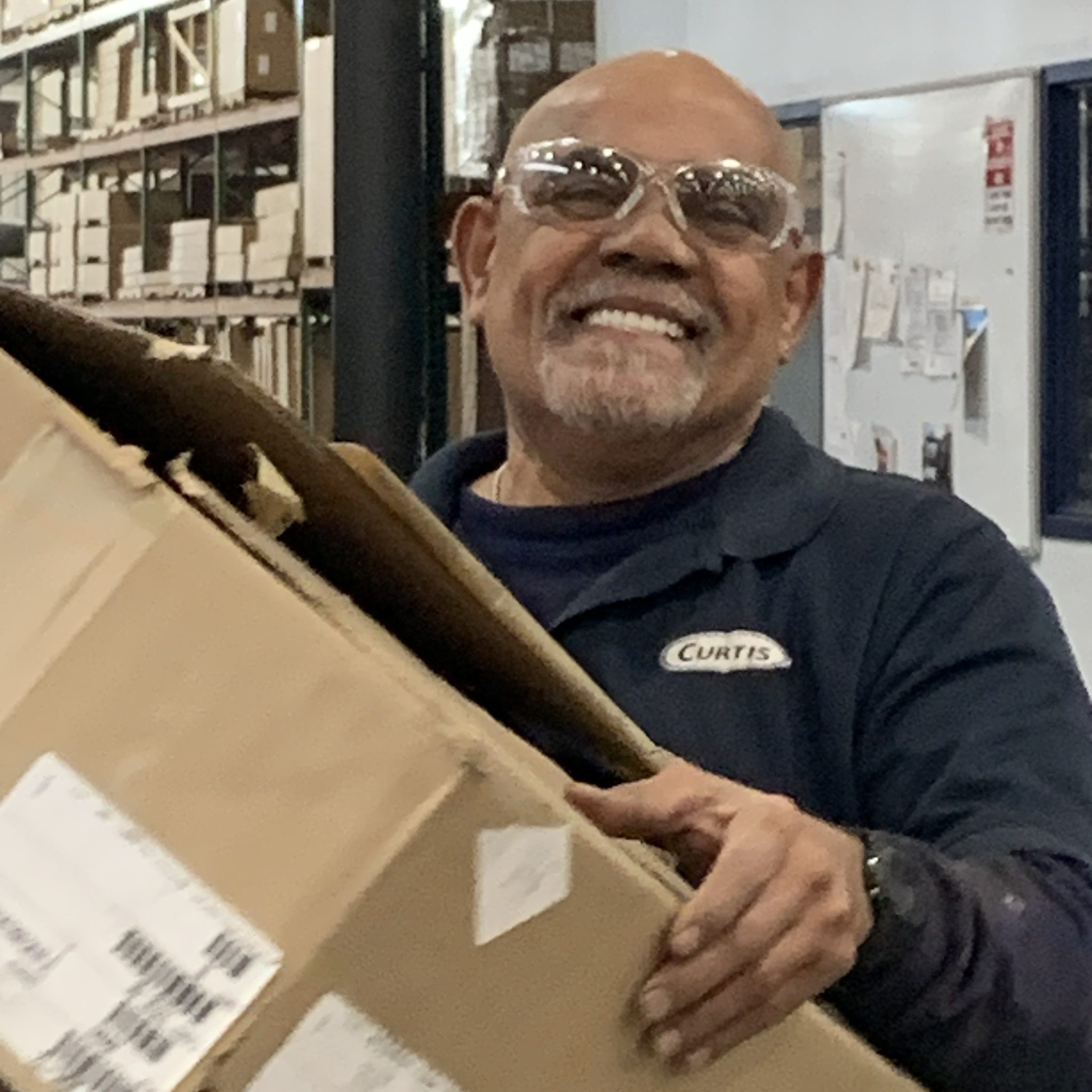 A smiling worker wearing clear safety glasses and a dark blue Curtis polo shirt holding a cardboard box in front of industrial shelving and a whiteboard.