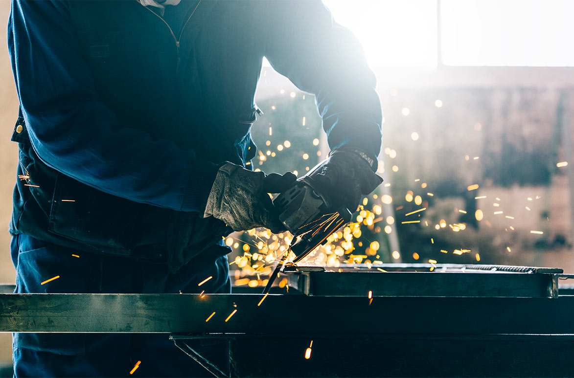 A worker in overalls and heavy-duty protective gloves operating a handheld angle grinder on a piece of metal.